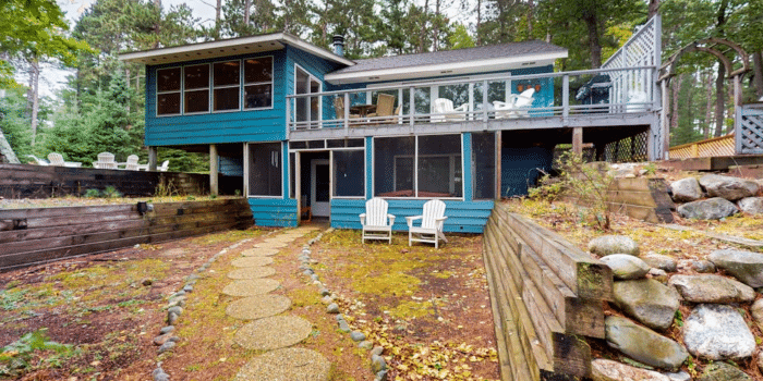 A two-story blue house in a forest with a second-floor deck, a screened-in porch on the lower level, and a stone path leading to two white chairs in the yard.
