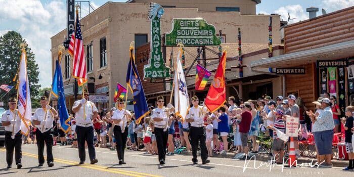 A parade with people holding flags marches down a street lined with spectators. Buildings with gift and souvenir signs are in the background.