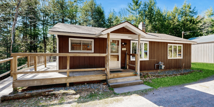 Single-story brown house with a wooden deck and porch in a wooded setting.