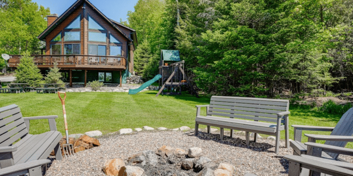 A backyard fire pit surrounded by benches, with a cabin and a wooden playset in the background.