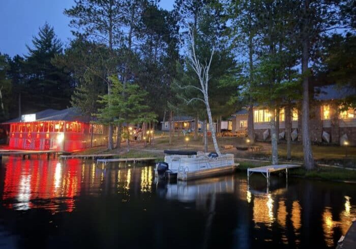 A lakeside scene at dusk shows a boat docked near illuminated buildings, including one with red lights reflecting on the water, surrounded by tall trees.