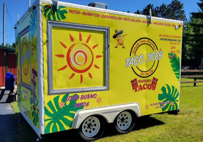 Bright yellow Taco Joe's food trailer with taco-themed decorations, sunlight graphic, menu items listed, and "Loco Bueno Tacos" text; parked outdoors on a sunny day.