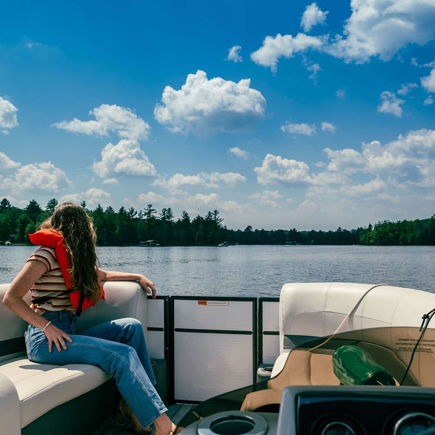 A person wearing a life jacket sits on a boat looking at the lake and surrounding trees under a partly cloudy sky.