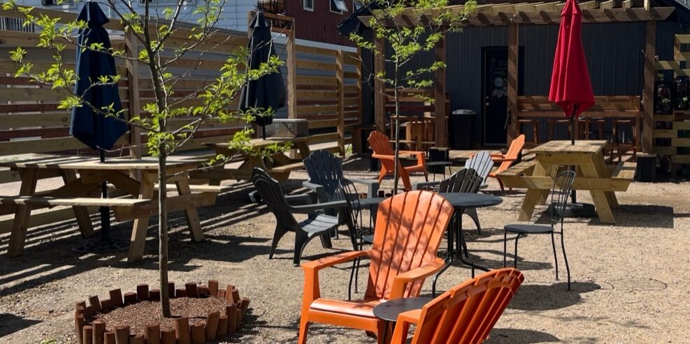 Outdoor seating area with orange and black chairs, picnic tables, and umbrella shades. Gravel ground and small green trees in the background under a partly cloudy sky.
