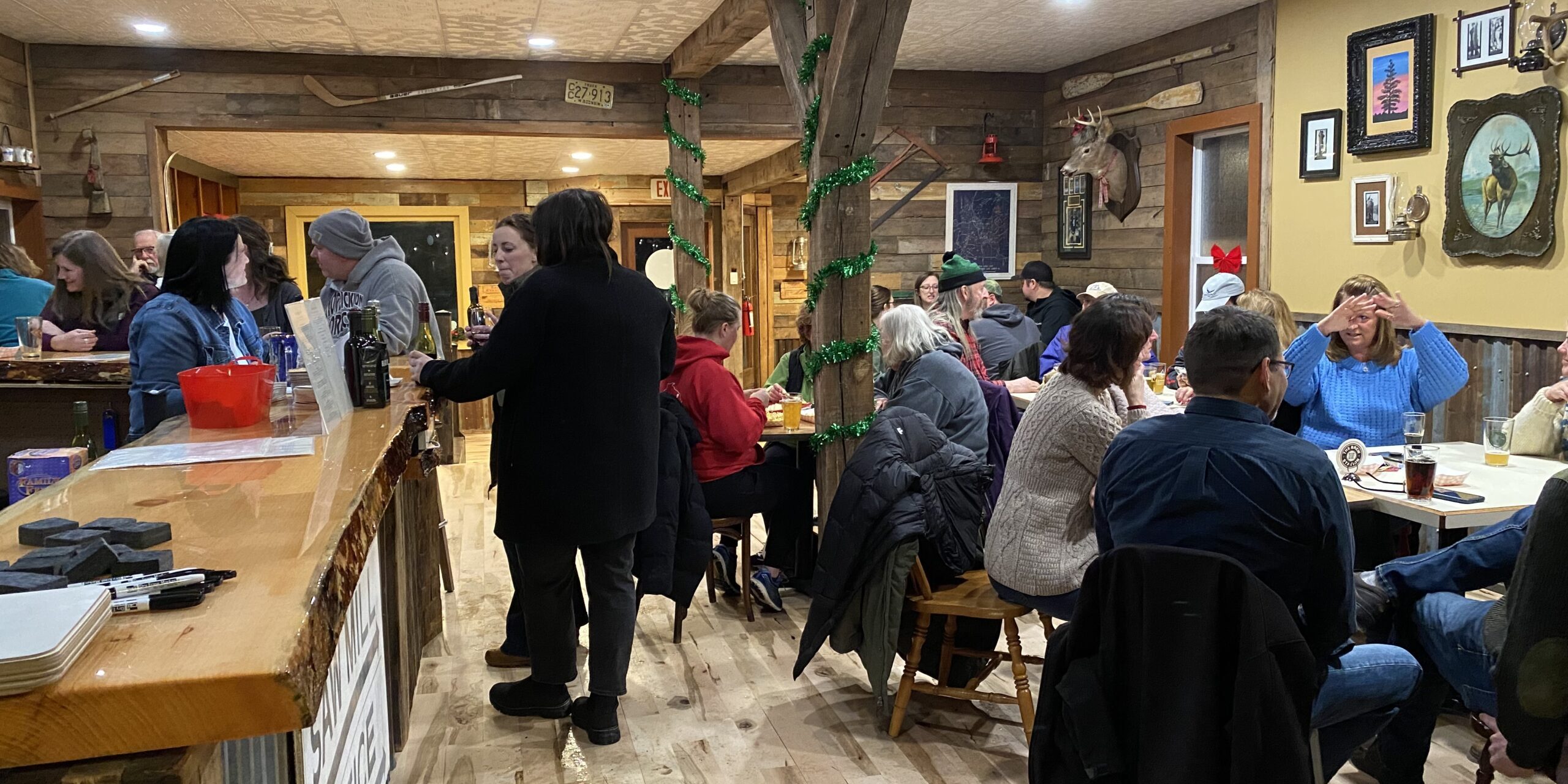 People dining in a cozy restaurant with wooden decor, seated at tables and a bar. Some are engaged in conversation, while others are eating or looking around.