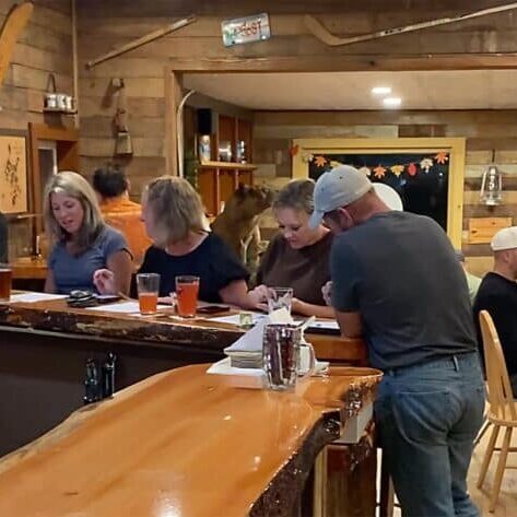 People sitting at a rustic wooden bar, drinking beer and looking at papers, while others sit and talk at nearby tables in a cozy, wood-paneled setting.