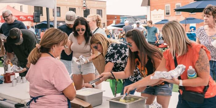 People serve themselves food from trays at an outdoor event. Some hold foil-wrapped items, while others gather around the table. The background shows more attendees and a brick building.