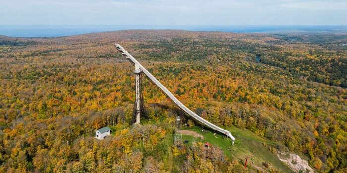 Aerial view of a large ski jump structure surrounded by a vast autumn forest landscape.