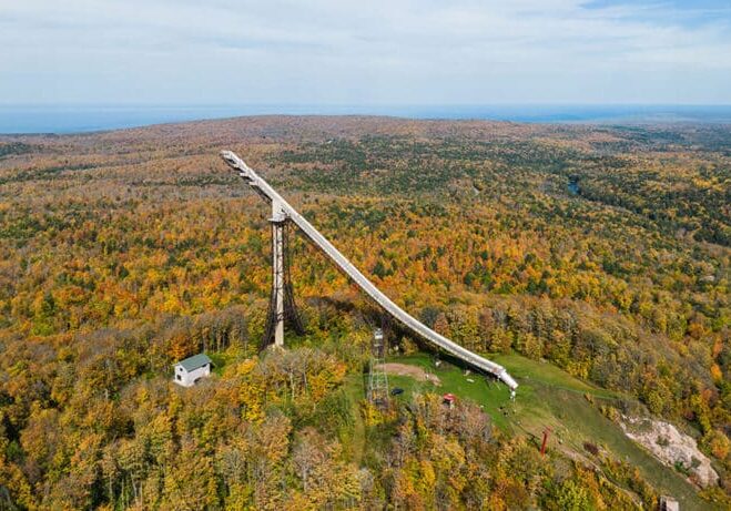 Aerial view of a large ski jump structure surrounded by a vast autumn forest landscape.