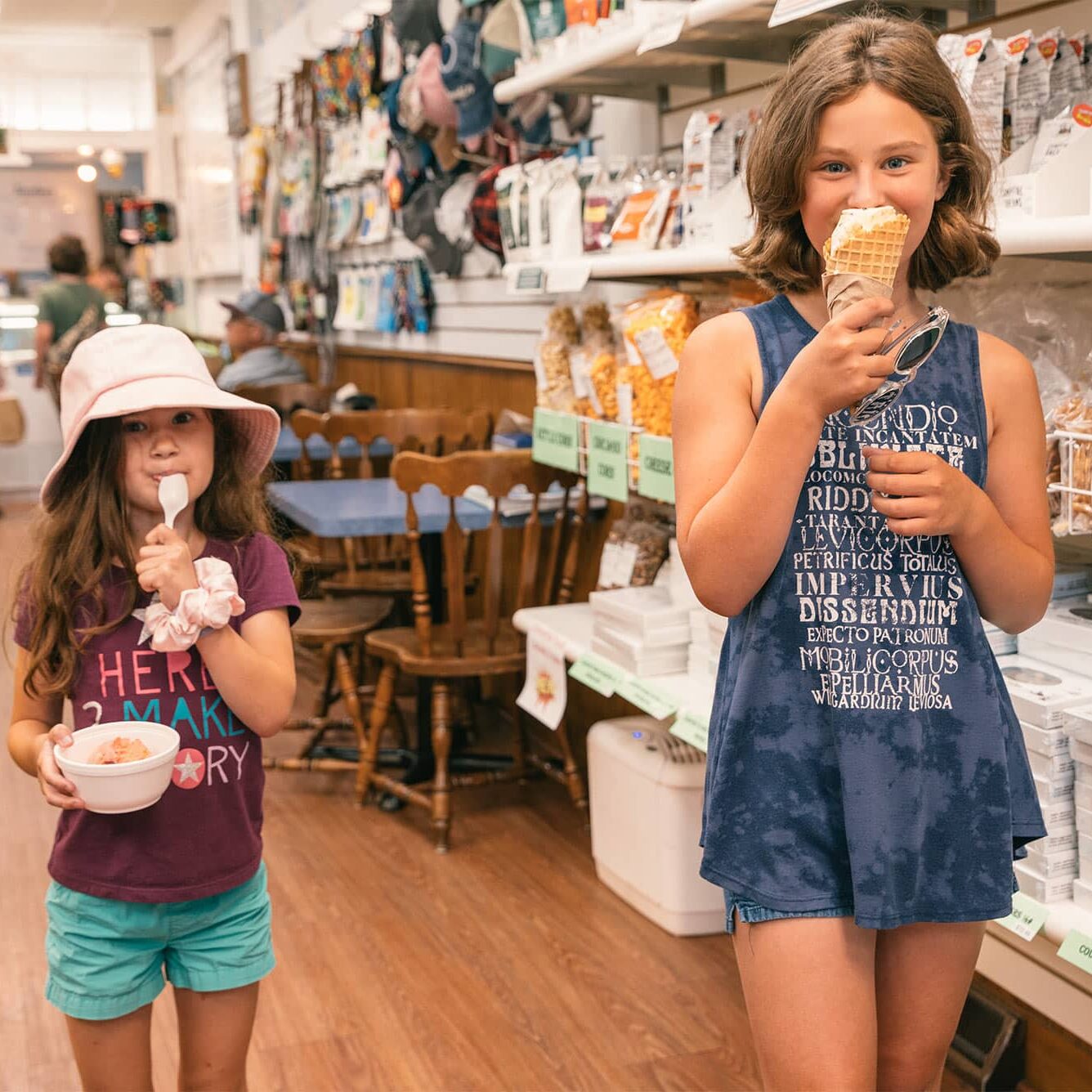 CountryStore Two young girls in a shop; one holds a bowl and spoon, the other eats an ice cream cone. Shelves of snacks and candy are in the background.