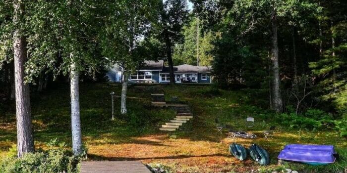 A lakeside cabin is partially hidden by trees, with a dock and steps leading up from the water. Canoes, chairs, and a blue floating mat are on the grassy shore.