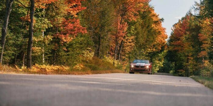 A red car drives along a rural road surrounded by trees with autumn foliage.