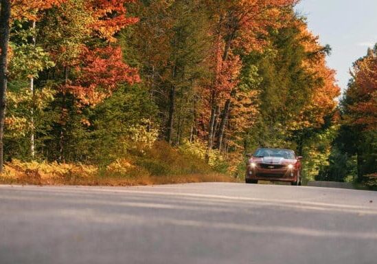 A red car drives along a rural road surrounded by trees with autumn foliage.