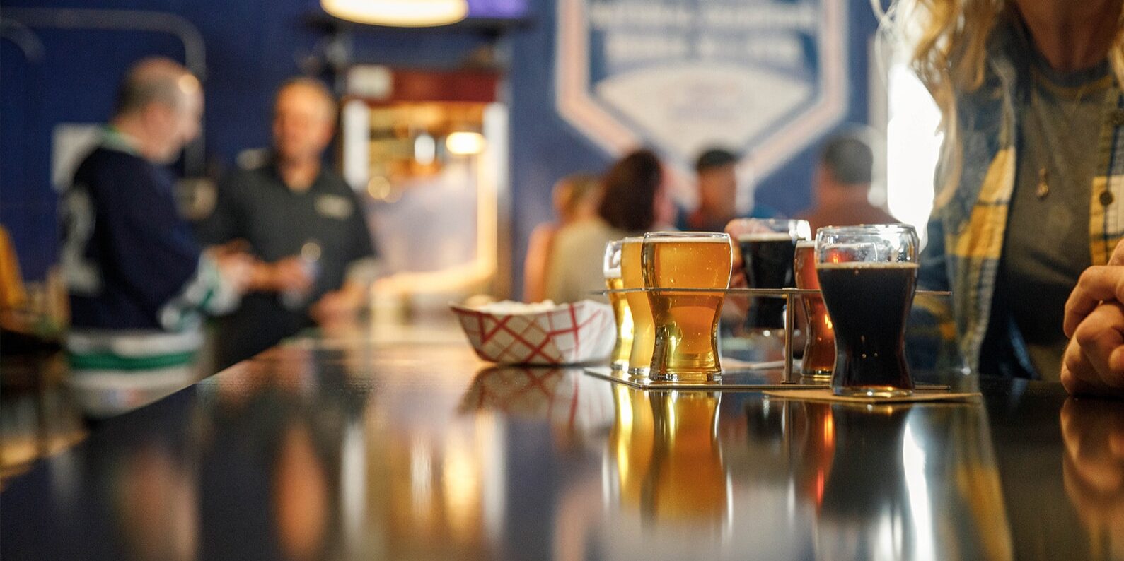 A variety of beers in glasses on a bar counter, with people in the background.