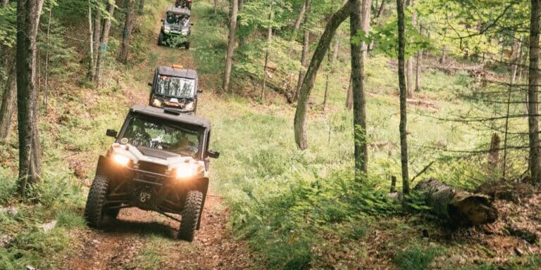 ER_ATV-UTV-2019 Three all-terrain vehicles drive up a dirt trail through a dense, green forest. The path is narrow and surrounded by tall trees and underbrush.