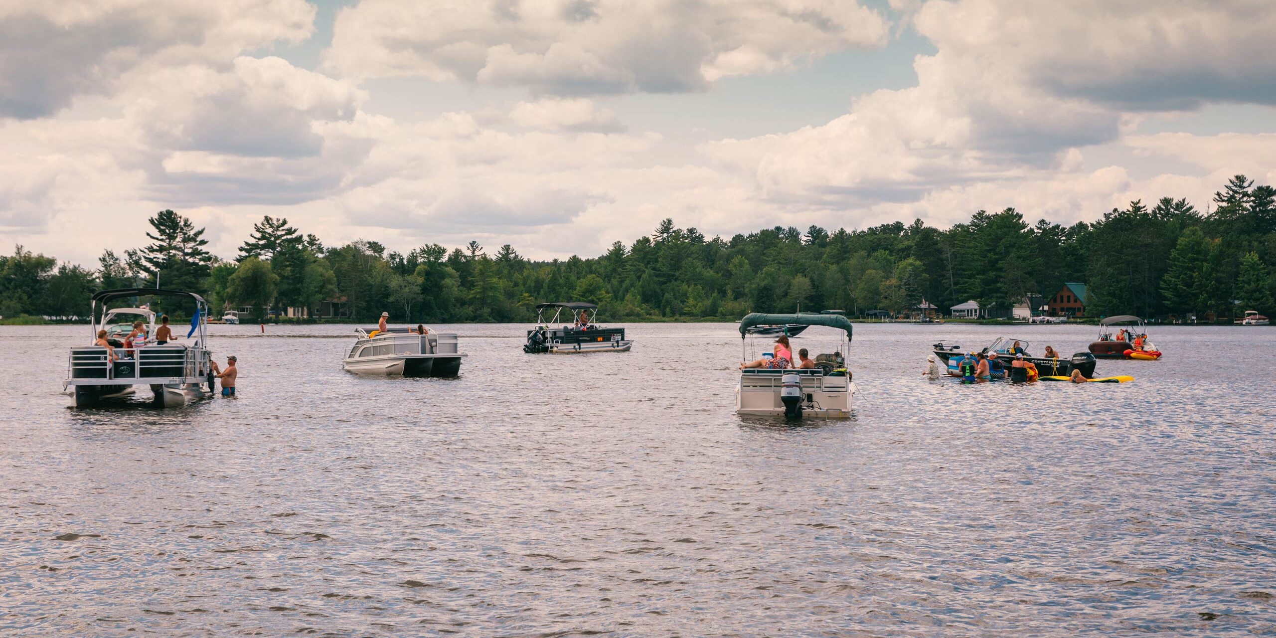 Several pontoon boats and people on kayaks are gathered on a lake under a partly cloudy sky, with trees and houses visible along the shoreline in the background.