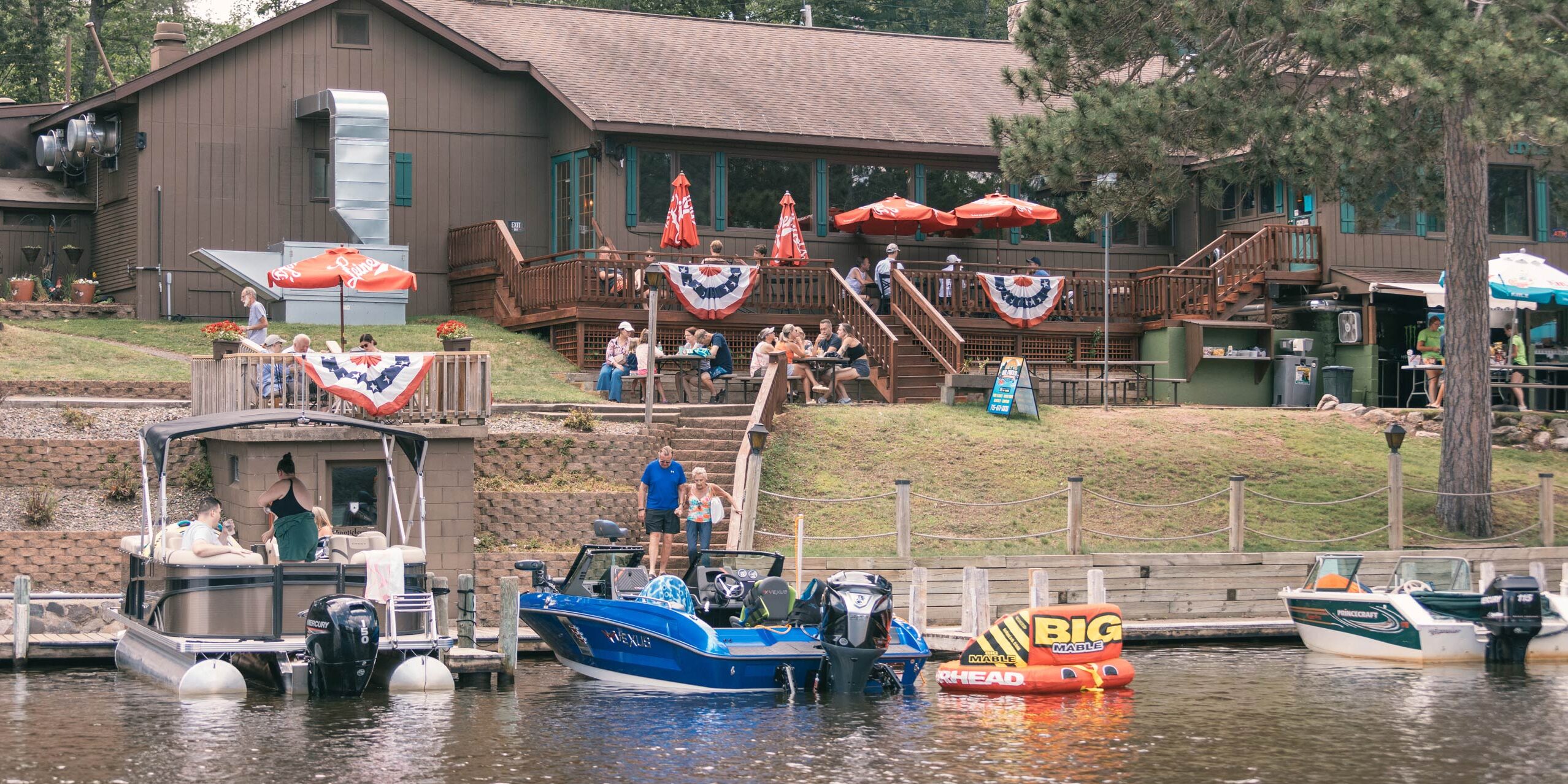 People gather outside a lakeside restaurant decorated with patriotic bunting, with several boats docked along the waterfront and a large "Big Red" tube visible.