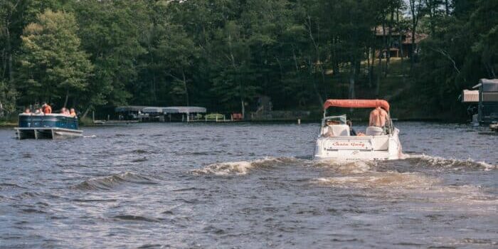 Two boats travel on a lake surrounded by trees, with one boat in the foreground labeled "Chasin' Gingers" and people visible on both boats.