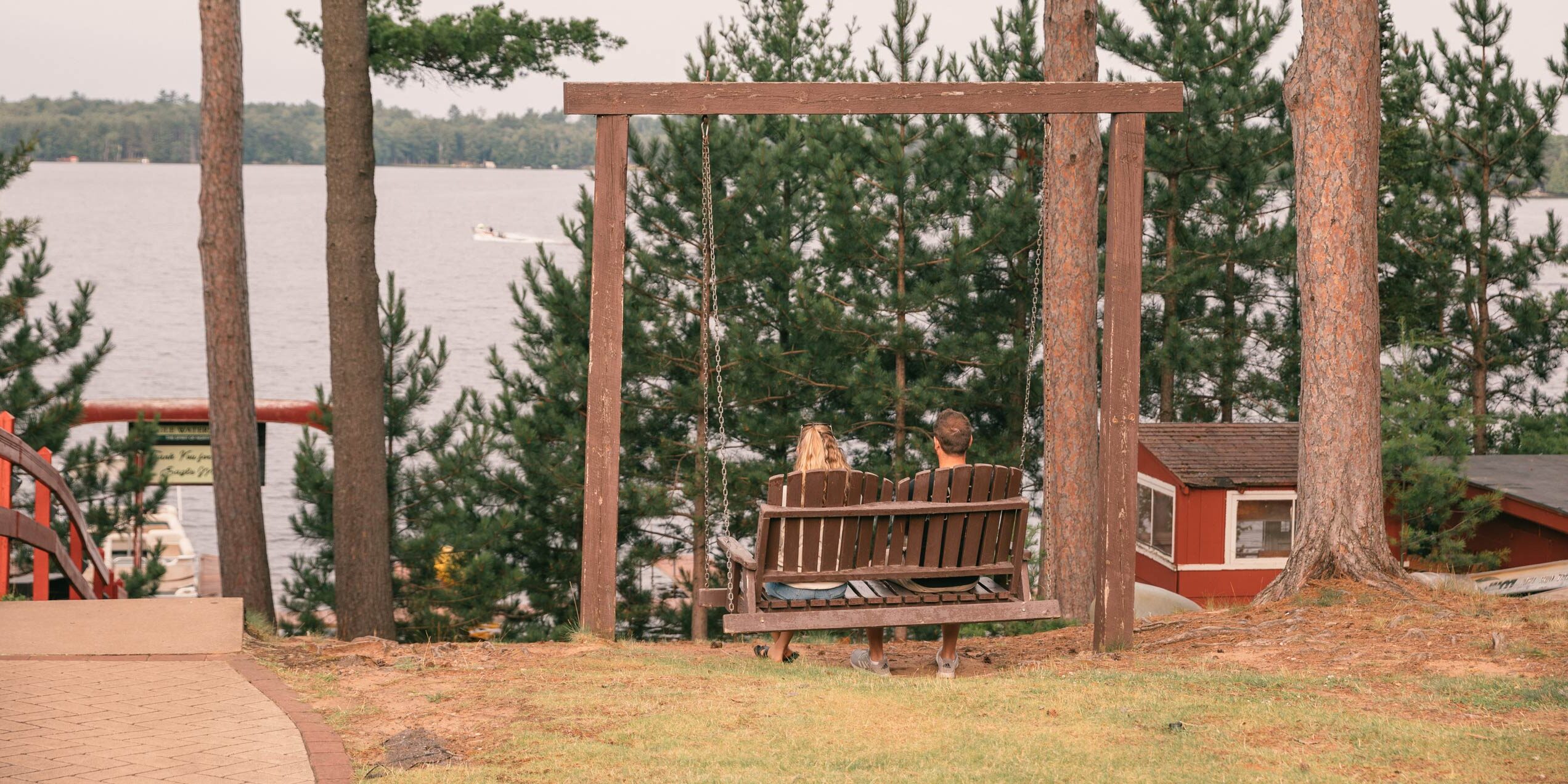 Two people sit on a wooden swing facing a lake, surrounded by trees and cabins, with a boat visible on the water in the background.