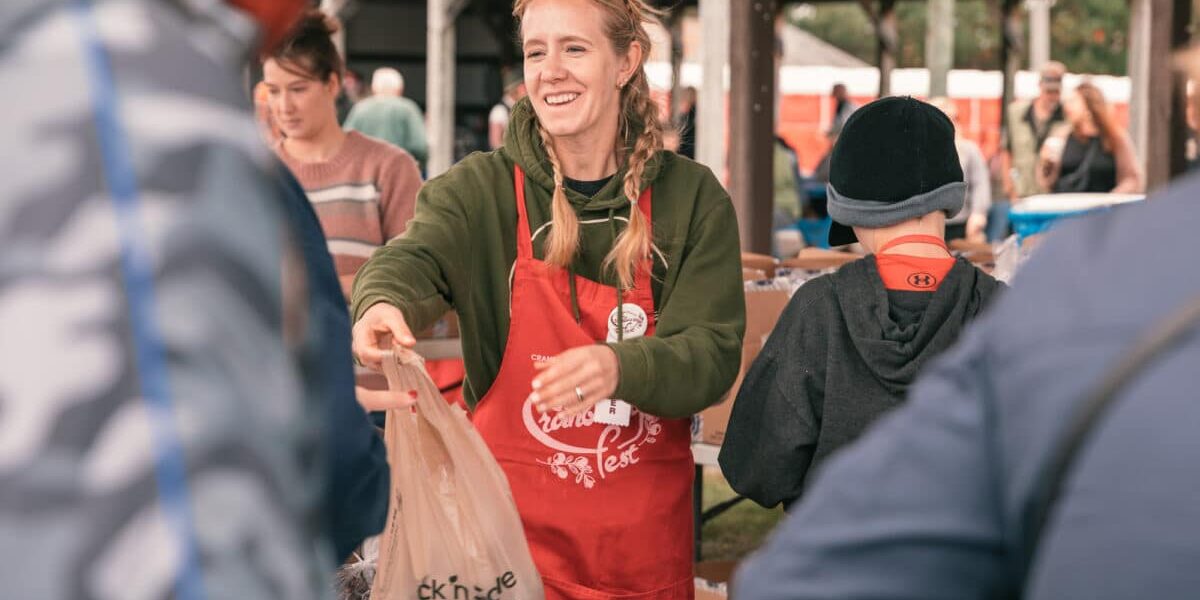 A woman wearing a red apron smiles as she hands a plastic bag to a person at an outdoor event or market. Other people are gathered around under a pavilion.
