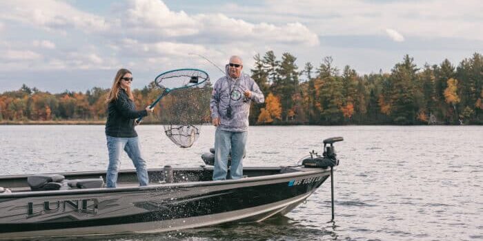 Two people on a fishing boat; one is holding a fishing net over the water while the other assists. Trees with autumn foliage and a cloudy sky are in the background.