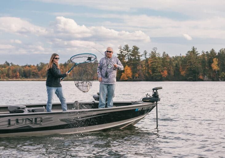 Two people on a fishing boat; one is holding a fishing net over the water while the other assists. Trees with autumn foliage and a cloudy sky are in the background.