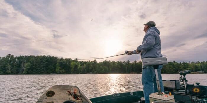 A person stands on a boat fishing with a rod on a lake under a cloudy sky, surrounded by trees and calm water.