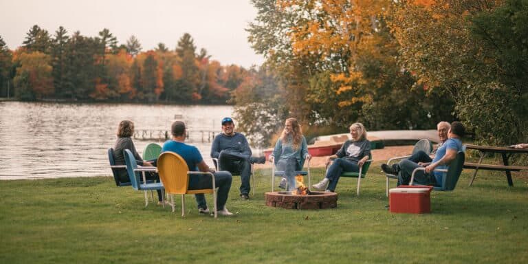 A group of people sit in chairs around a fire pit by a lake on a grassy area with autumn trees in the background.