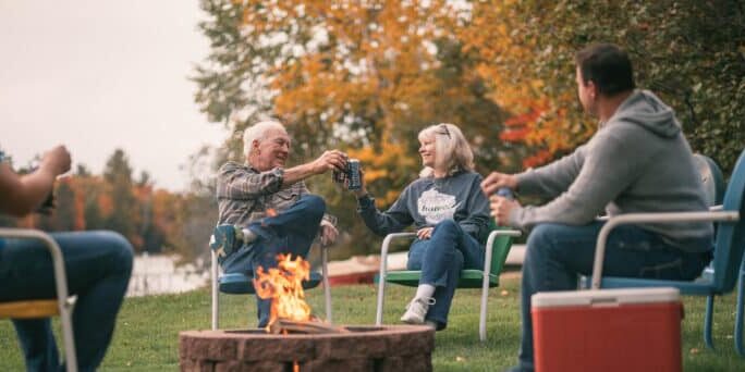 Four people sit around a fire pit outdoors on a grassy lawn, holding drinks and chatting, with autumn trees and a body of water in the background.