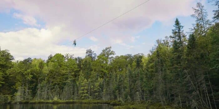 Person zip-lining over a forested lake under a cloudy sky.