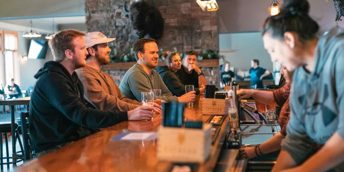 A group of people sitting at a wooden bar counter with drinks, while a bartender serves them. A stone wall and various decorations are in the background.