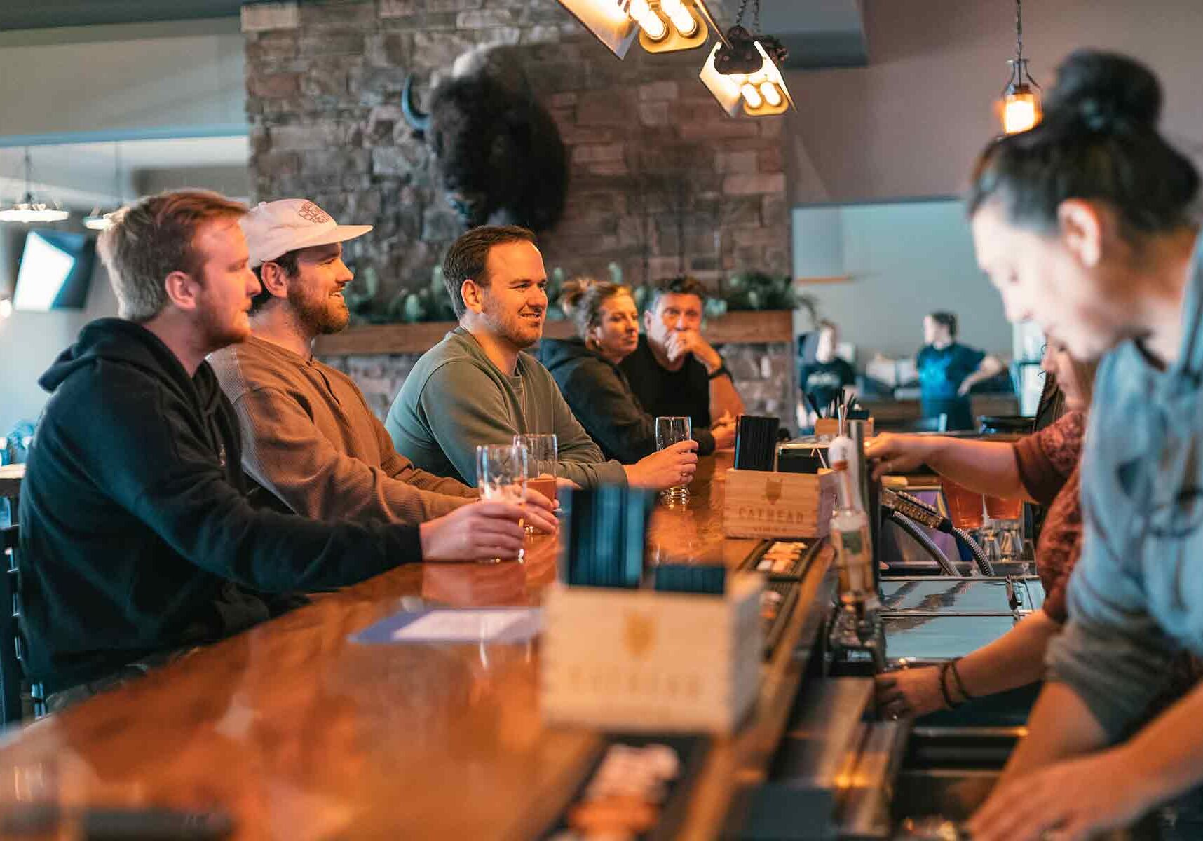 A group of people sitting at a wooden bar counter with drinks, while a bartender serves them. A stone wall and various decorations are in the background.