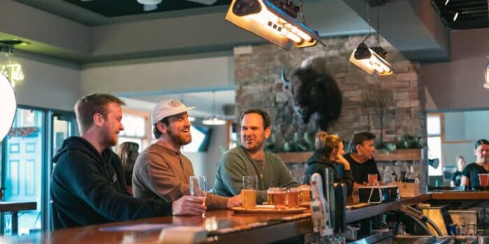 Three men sit at a bar with drinks, talking and smiling; other patrons are in the background, and a buffalo head is mounted on the wall.