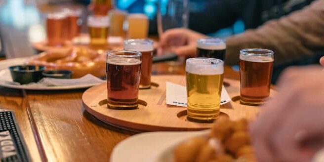 A wooden tray with four small glasses of beer sits on a bar table, surrounded by plates of food and people’s hands reaching for drinks.