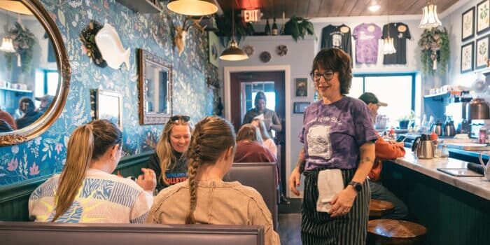 Server smiling and standing near a table with seated customers in a cozy, decorated diner.