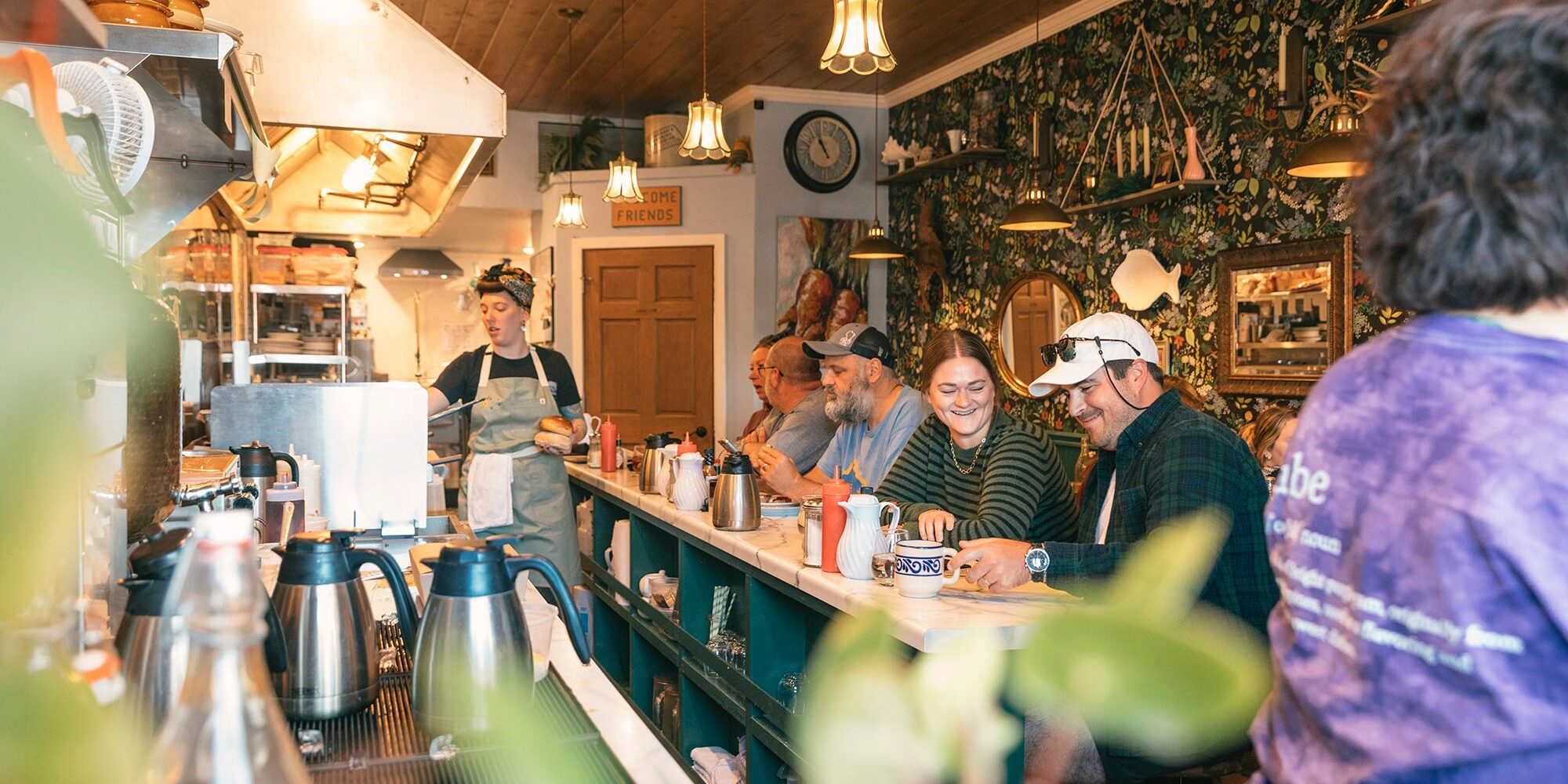 A group of people sit at a counter inside a cozy diner. A staff member in an apron works behind the counter. The ambiance is warm with decorative wallpaper and overhead lighting.