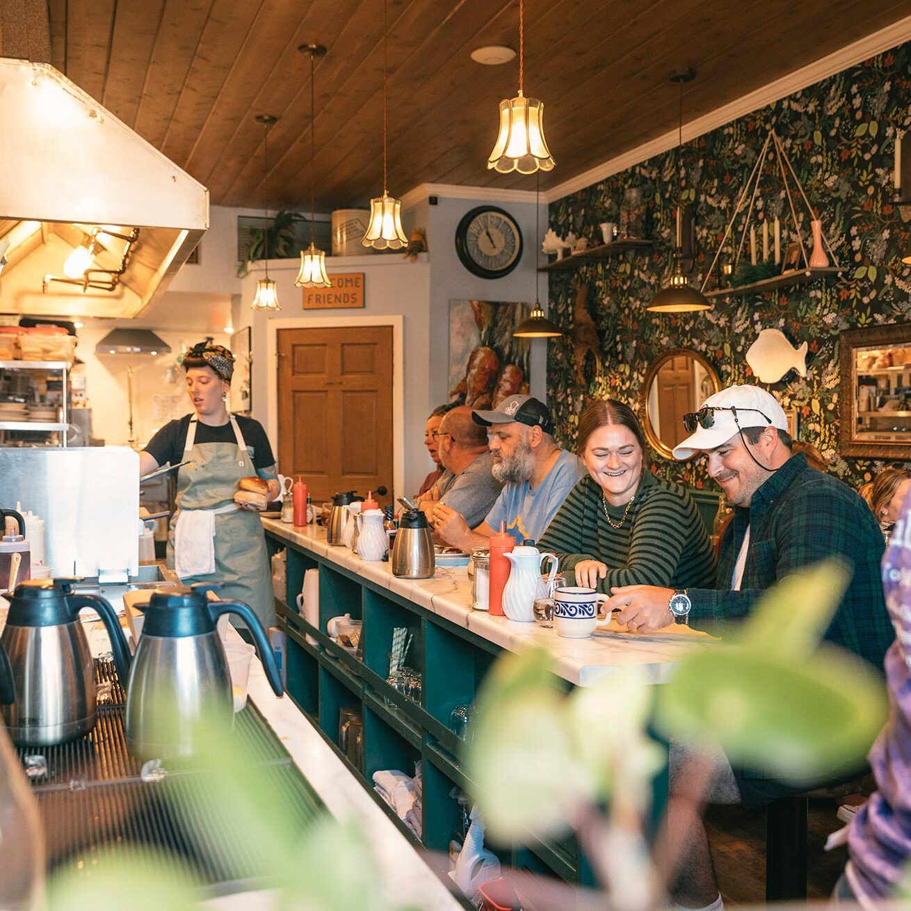 ER_Fall_Toad_In_The_Hole_2024 A group of people sit at a counter inside a cozy diner. A staff member in an apron works behind the counter. The ambiance is warm with decorative wallpaper and overhead lighting.