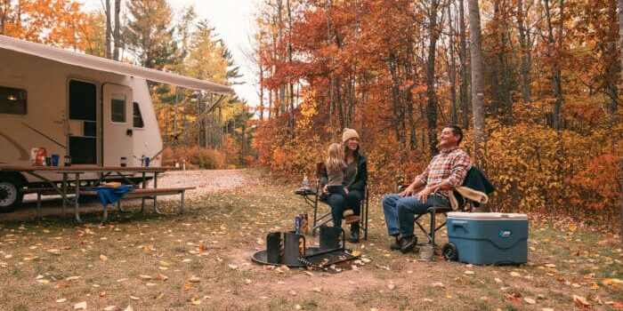 A man, woman, and child sit around a fire pit at a campsite with an RV and picnic table nearby, surrounded by autumn foliage.