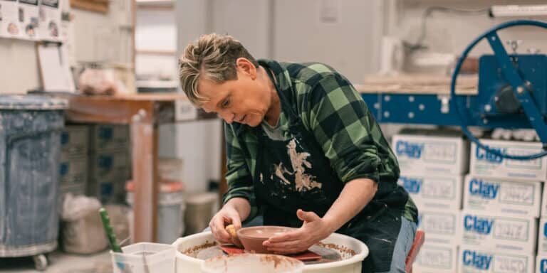 ER_Fall_Warehouse_Arts_2024 A person wearing a flannel shirt shapes clay on a pottery wheel in a workshop filled with pottery tools and materials.