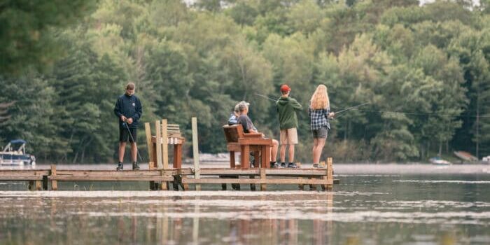 Four people are fishing from a wooden dock on a calm lake, surrounded by trees. Two stand with fishing rods, one is seated, and one watches nearby.