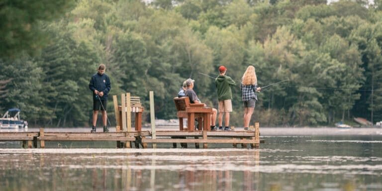 Four people are fishing from a wooden dock on a calm lake, surrounded by trees. Two stand with fishing rods, one is seated, and one watches nearby.