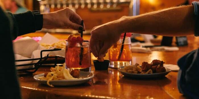 Two people at a bar table with cocktails and plates of fried food.