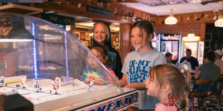 ER_LPs_Pizza_and_Pasta Four children play a bubble hockey arcade game in a lively, indoor setting with tables and people in the background.