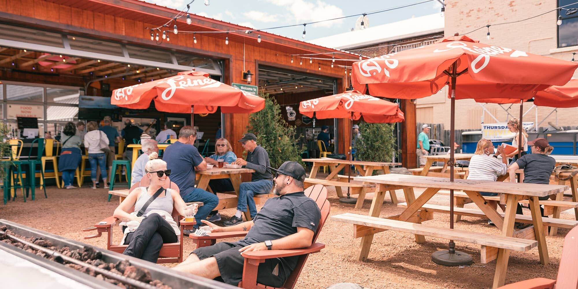 People sit at picnic tables and wooden chairs under red umbrellas at an outdoor bar or restaurant on a sunny day.