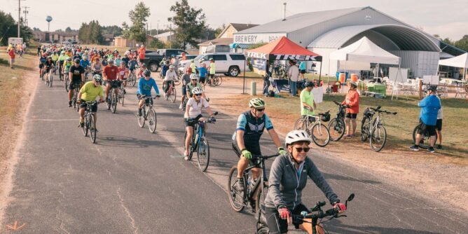 A group of cyclists participates in a biking event on a country road, passing by a brewery and a Trek tent.