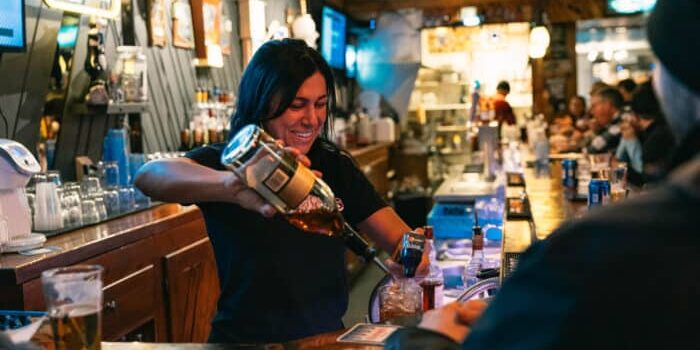 A bartender pours liquor into a glass behind a bar in a busy, warmly lit establishment with customers seated.