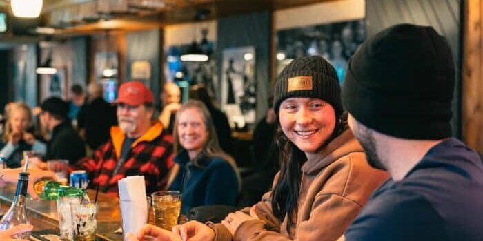 People sit at a bar counter, drinking and talking. A woman in a beanie smiles at a man beside her. The atmosphere is casual and friendly with wood paneling and dim lighting.