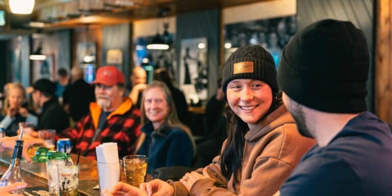 People sit at a bar counter, drinking and talking. A woman in a beanie smiles at a man beside her. The atmosphere is casual and friendly with wood paneling and dim lighting.