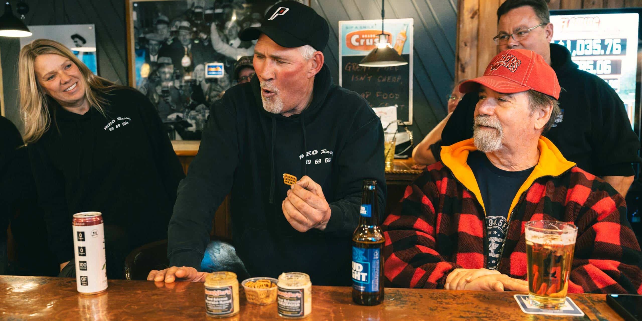 A group of people react as a man in a black hoodie eats something at a bar counter, with drinks and small food containers in front of them.