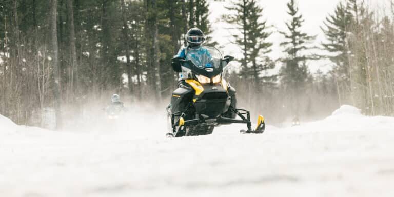 Person riding a yellow snowmobile on a snowy trail through a forest, with other snowmobiles visible in the background.